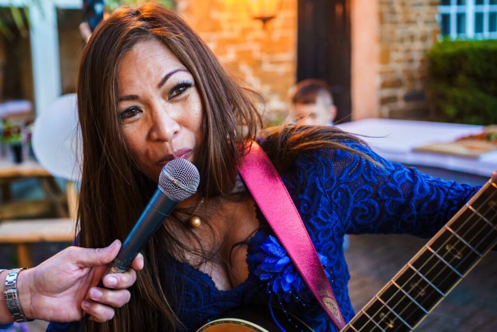 A woman with long hair, dressed in a blue top, is singing into a microphone held by another person. She appears to be playing a guitar, indicated by the visible guitar strap over her shoulder. The setting looks like an outdoor courtyard with brick walls and a wooden table in the background. Image by Documentary Wedding Photographer Lewis Bishop Photography.
