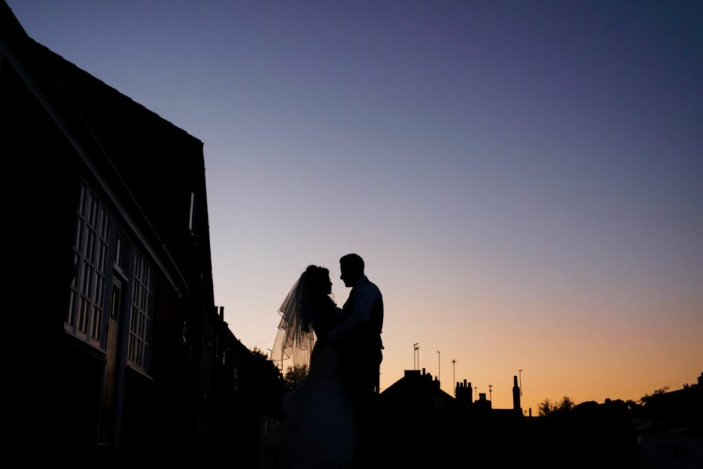 Silhouetted against a twilight sky, a bride and groom share an intimate moment. The dusky hues of the sunset create a romantic backdrop, highlighting the outlines of buildings and chimneys in the distance. Image by Documentary Wedding Photographer Lewis Bishop Photography.