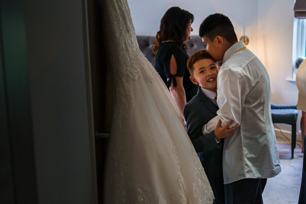 Two young boys, one in a white shirt and the other in a suit, hug and smile warmly in a dimly lit room. A woman with dark hair stands in the background beside a white wedding dress hanging near a doorframe. The mood is intimate and joyful. Image by Documentary Wedding Photographer Lewis Bishop Photography.