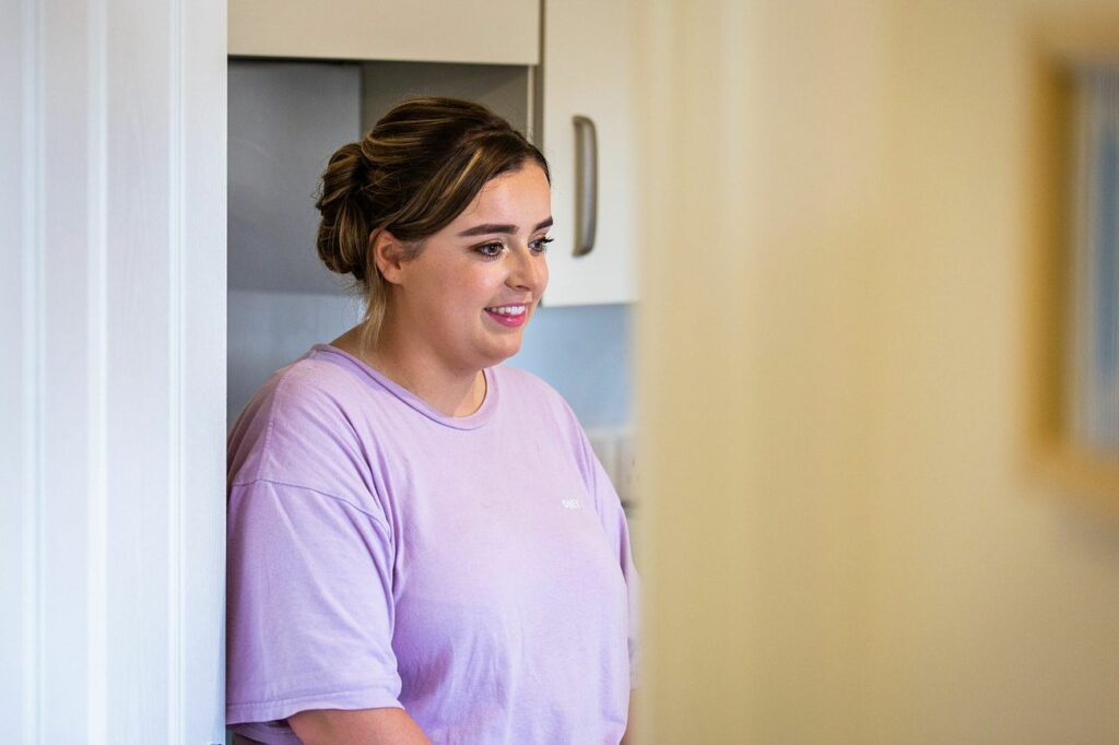 A woman with tied-back hair and a light purple shirt stands in a room, leaning against a doorframe. She smiles gently, looking towards something or someone off-camera. The background shows part of a kitchen with white cabinets and a partial view of a framed picture. Image by Documentary Wedding Photographer Lewis Bishop Photography.