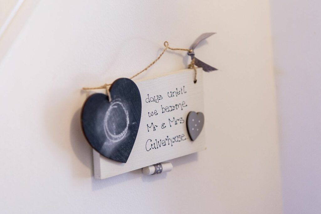 A small white sign hangs on a wall, adorned with a chalkboard heart and a polka-dotted heart. The text on the sign reads, "days until we become Mr & Mrs Culverhouse." A piece of twine holds a small knotted ribbon above the sign. Image by Documentary Wedding Photographer Lewis Bishop Photography.