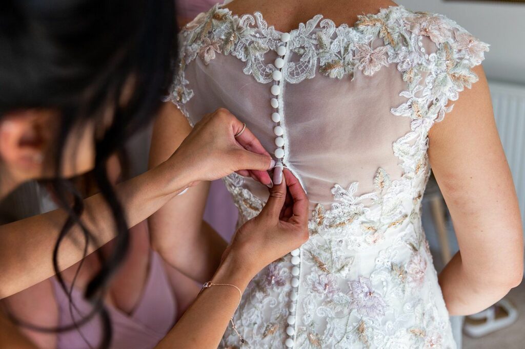Close-up of a bride's back as her dress is being buttoned. The lace dress features intricate floral details and a row of small, white buttons. Two hands are visible, assisting with fastening the gown. The bride's dark hair is partly visible on the left. Image by Documentary Wedding Photographer Lewis Bishop Photography.