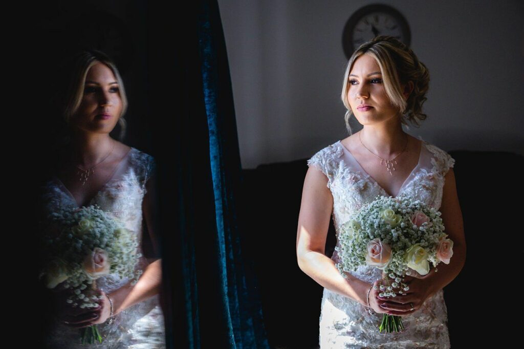 A bride in a white lace gown holds a bouquet of white and pale pink flowers. She stands near a window with natural light illuminating her, and her reflection is visible in a dark mirror to her left. The background is dimly lit, with a clock faintly visible. Image by Documentary Wedding Photographer Lewis Bishop Photography.