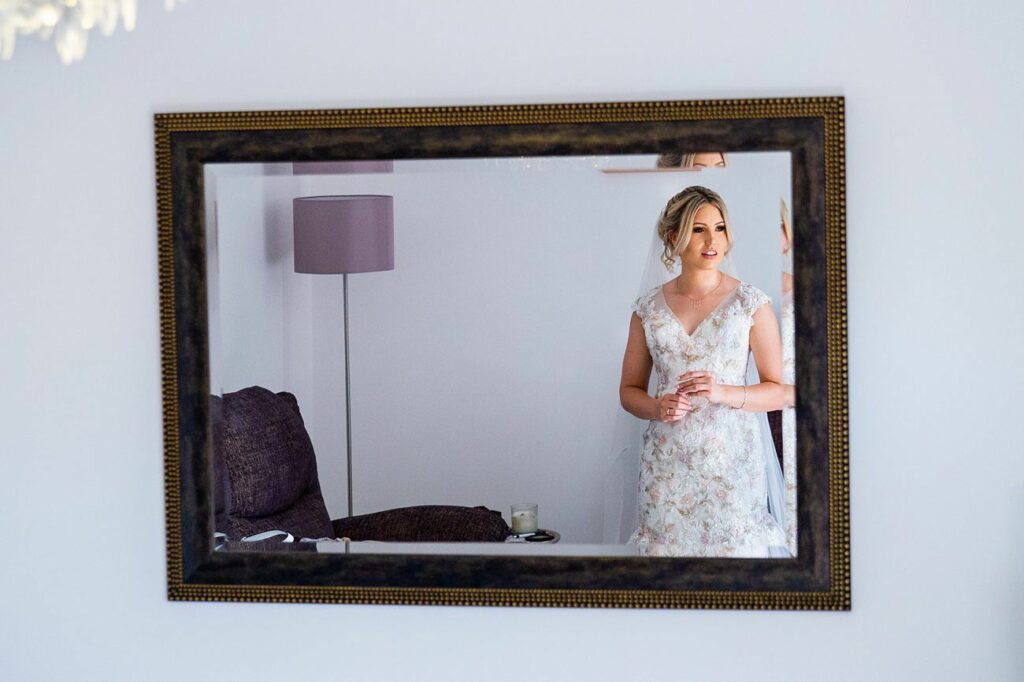 A bride in a white floral wedding dress is reflected in a large, ornate framed mirror. She stands in a light-colored room with a purple lampshade and a candle on a small table. Her hands are clasped, and she looks content and thoughtful. Image by Documentary Wedding Photographer Lewis Bishop Photography.