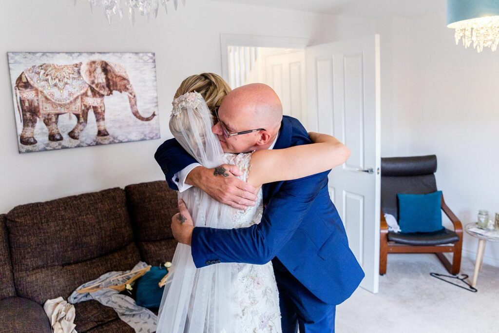 A person in a wedding dress is embracing another individual in a blue suit inside a room. A couch is seen to the left, with clothes lying on it. On the white wall behind them is an elephant painting. A chandelier and a chair with a cushion are also visible. Image by Documentary Wedding Photographer Lewis Bishop Photography.