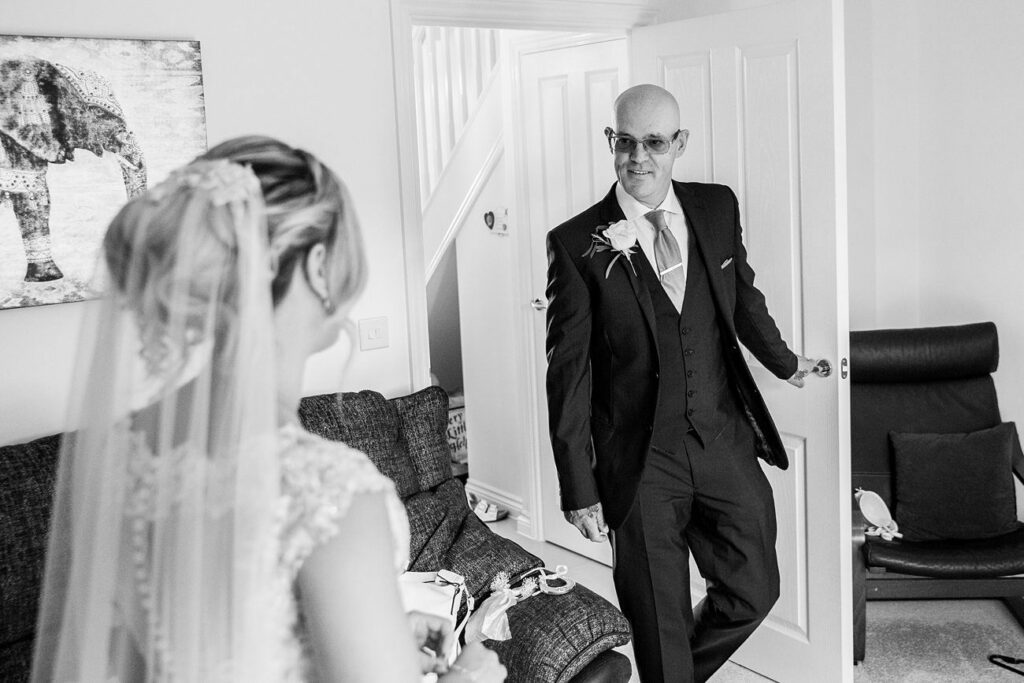 A person in a wedding dress, presumably the bride, stands facing a man in a suit, who is smiling as he enters the room. The room has a couch, a piece of artwork on the wall, and an open door. The scene appears emotional and celebratory. Image by Documentary Wedding Photographer Lewis Bishop Photography.
