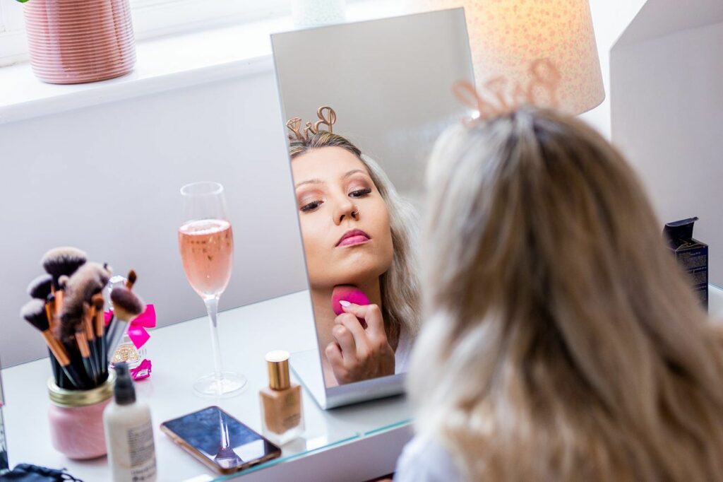 A person with long blonde hair is applying makeup using a pink sponge while looking into a mirror. On the table are various makeup brushes, cosmetics, a smartphone, and a glass of pink champagne. A lamp and a plant pot are in the background. Image by Documentary Wedding Photographer Lewis Bishop Photography.