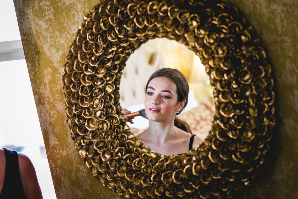 A woman is reflected in a round, ornate mirror as she applies makeup with a brush. The mirror has a circular frame made up of small, gold, flower-like designs. The woman has a focused expression and is wearing a black tank top. Image by Documentary Wedding Photographer Lewis Bishop Photography.