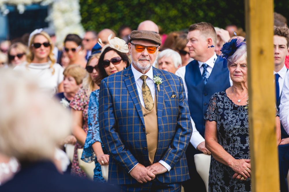 An older man wearing a plaid suit, flat cap, and orange-tinted sunglasses stands among a crowd at an outdoor event. He is flanked by a woman in a floral dress on his right. The crowd, dressed in formal attire, is focused on something happening in front of them. Image by Documentary Wedding Photographer Lewis Bishop Photography.