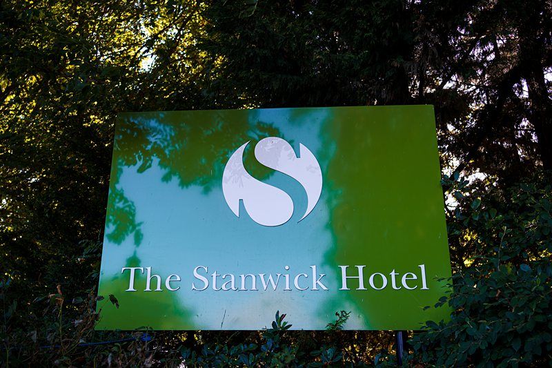 A green sign with the white logo of an 'S' shape and text reading "The Stanwick Hotel" stands amidst greenery, with trees in the background reflecting sunlight. Image by Documentary Wedding Photographer Lewis Bishop Photography.