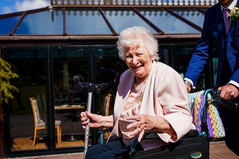 An elderly woman with white hair sits in a wheelchair, smiling and holding a walking stick. She wears a pink jacket. Behind her is a person in a suit pushing the wheelchair. They are outdoors in front of a glass building with a blue sky above. Image by Documentary Wedding Photographer Lewis Bishop Photography.