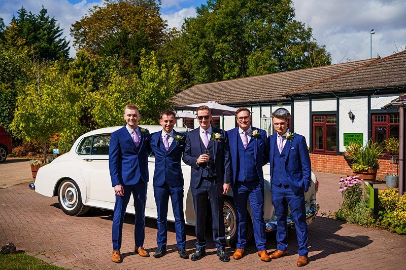 Five men dressed in matching navy blue suits with pink ties stand in front of a vintage white car outside a building. One man in the center holds a drink, and they are all smiling at the camera. Trees and bushes can be seen in the background. Image by Documentary Wedding Photographer Lewis Bishop Photography.