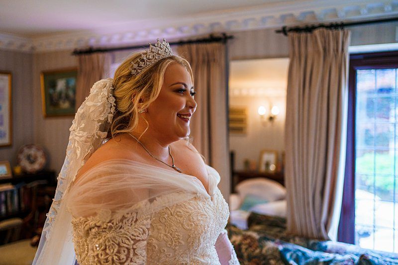 A smiling bride in a beautiful white wedding dress with lace detailing and a long veil stands indoors near a window with curtains. She is wearing a sparkling tiara and jewelry, looking happy and radiant. The room has elegant decor with framed art on the walls. Image by Documentary Wedding Photographer Lewis Bishop Photography.