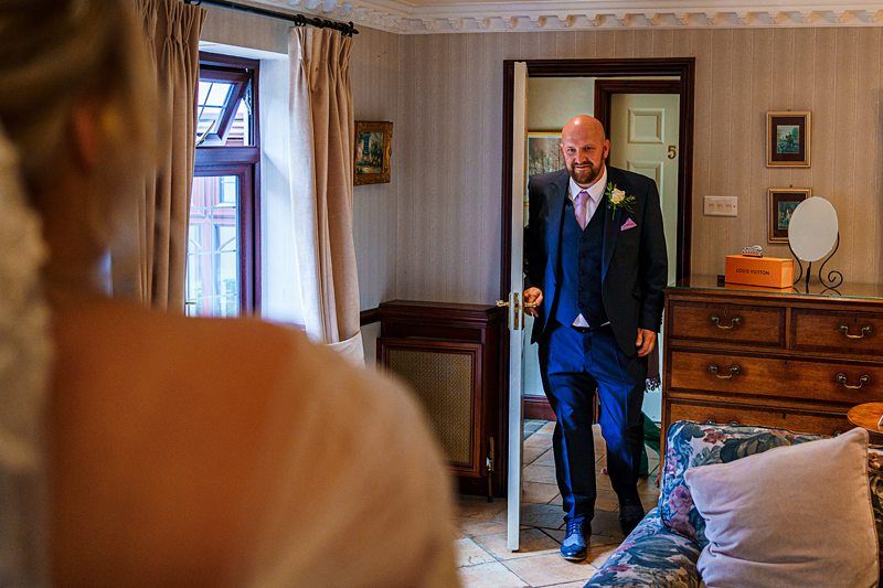 A man in a suit smiles as he enters a room, looking towards a woman in a wedding dress with her back to the camera. The room has a dresser with a mirror, a floral-patterned sofa, and light-colored walls adorned with framed pictures. Image by Documentary Wedding Photographer Lewis Bishop Photography.