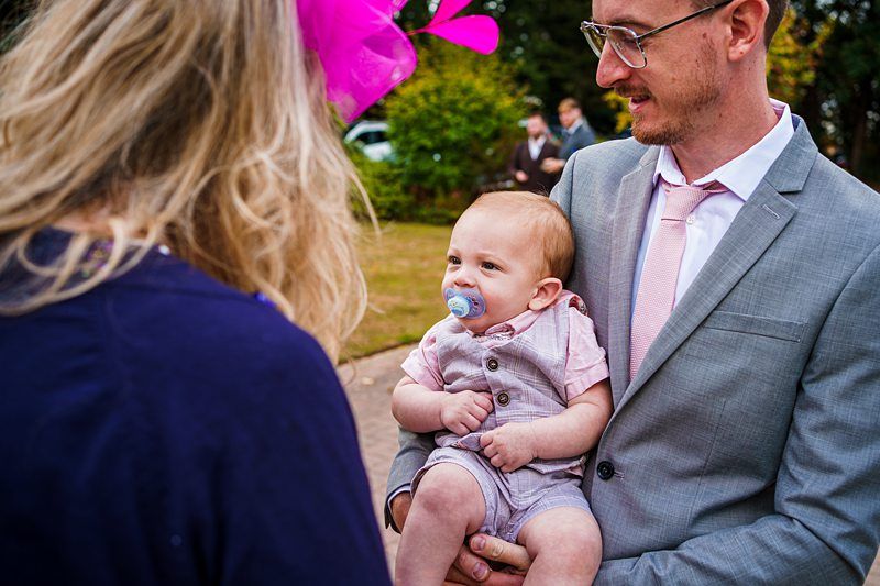 A man in a gray suit and pink tie holds a baby with a pacifier in his mouth. The man smiles at a woman with long blonde hair and a pink fascinator, seen from behind. They are outdoors with greenery in the background. Image by Documentary Wedding Photographer Lewis Bishop Photography.