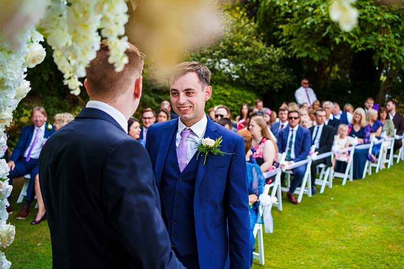 Two grooms stand facing each other and smiling during an outdoor wedding ceremony. Guests in formal attire are seated on white chairs in the background, attentively watching the couple. The setting is a lush, green garden with decorative flowers visible in the foreground. Image by Documentary Wedding Photographer Lewis Bishop Photography.