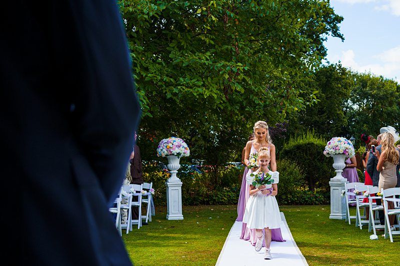 A young flower girl in a white dress and a woman in a lavender gown walk down an outdoor aisle adorned with white chairs and flower arrangements. The scene is set in a garden with greenery and trees, under a partly cloudy sky. Image by Documentary Wedding Photographer Lewis Bishop Photography.