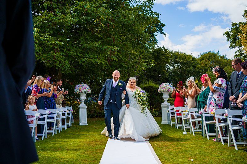 A bride and groom joyfully walk down the aisle outdoors. The groom is dressed in a dark suit, and the bride wears a white wedding gown, holding a large bouquet. Guests on both sides of the aisle applaud and take photos. The setting is lush with green trees and flowers. Image by Documentary Wedding Photographer Lewis Bishop Photography.