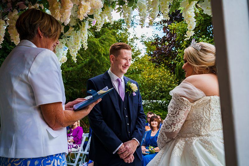 A bride and groom, dressed formally, stand facing each other under a floral arch during their outdoor wedding ceremony. An officiant in a white blazer and floral skirt holds a book and speaks, while guests watch in the background amidst lush greenery. Image by Documentary Wedding Photographer Lewis Bishop Photography.