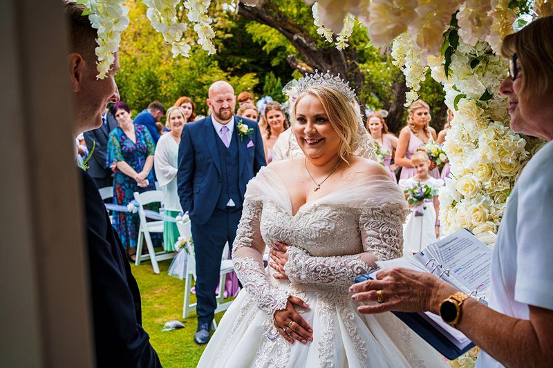 A bride in a white lace wedding dress and tiara smiles radiantly at the groom, standing under a floral archway during an outdoor ceremony. A group of guests, including bridesmaids in purple dresses, look on joyfully from the background. The officiant holds an open book. Image by Documentary Wedding Photographer Lewis Bishop Photography.