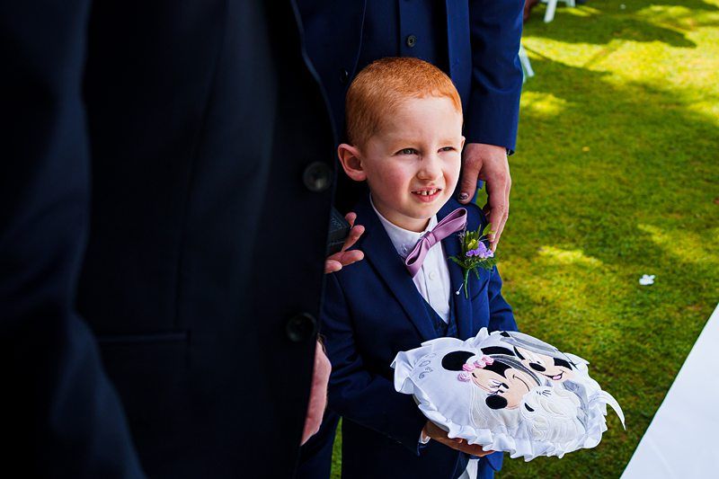 A young boy with red hair stands outside on a grassy area, wearing a navy suit with a bow tie and boutonniere. He holds a decorative pillow featuring a cartoon mouse couple. An adult's hand rests on his shoulder, and other adults in suits are nearby. Image by Documentary Wedding Photographer Lewis Bishop Photography.