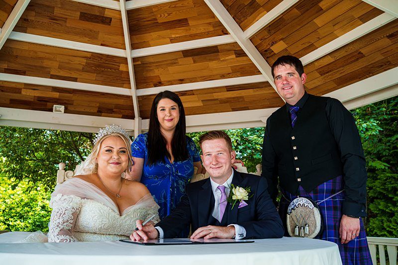 A bride in a white dress and tiara sits beside a groom in a black suit signing a document at an outdoor wedding ceremony. They are joined by a woman in a blue dress and a man wearing a kilt. They are all under a wooden gazebo with greenery in the background. Image by Documentary Wedding Photographer Lewis Bishop Photography.