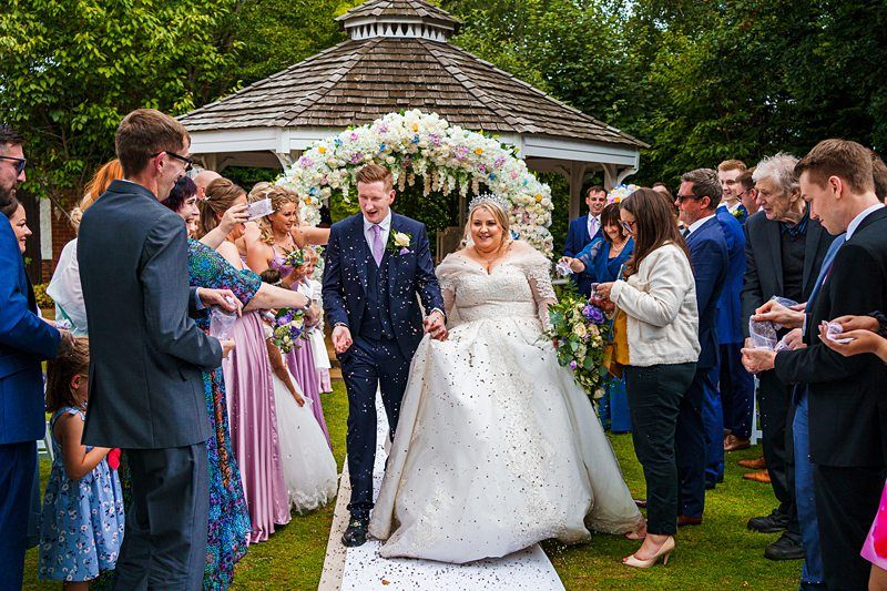 A newly married couple, dressed in wedding attire, walks down an outdoor aisle beneath a floral arch. The bride, in a white gown, and the groom, in a suit, are surrounded by guests in colorful outfits who are throwing confetti. A small gazebo is visible in the background. Image by Documentary Wedding Photographer Lewis Bishop Photography.