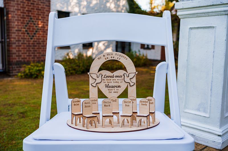 A memorial display on a white chair features a central plaque with doves and an inscription for a lost loved one. Smaller wooden chairs, each labeled with names like "Grandma Helene" and "Uncle Jack," symbolize absent family members. Image by Documentary Wedding Photographer Lewis Bishop Photography.