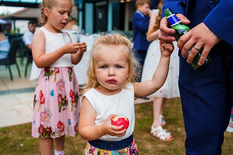 A blonde toddler in a white top and colorful floral skirt holds an apple and the hand of an adult in a blue suit. Two other children are in the background, one playing with a small object and the other holding a paper streamer. The setting appears to be outdoors. Image by Documentary Wedding Photographer Lewis Bishop Photography.
