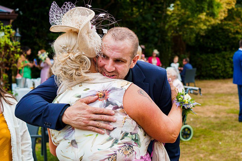 A man in a navy suit warmly embraces a woman in a floral dress at an outdoor gathering. The woman is wearing a beige fascinator with decorative elements. The background features other people and greenery, suggesting a celebratory event like a wedding or garden party. Image by Documentary Wedding Photographer Lewis Bishop Photography.