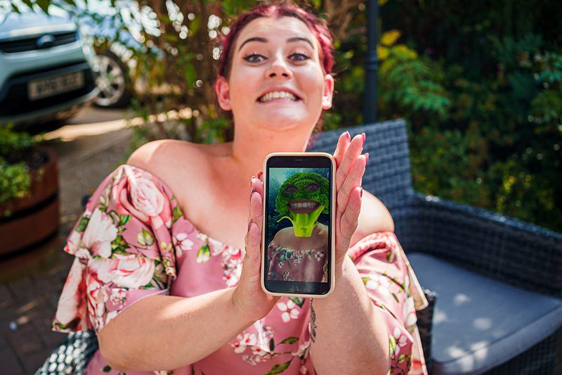 A person with a cheerful expression holds up a smartphone displaying a humorous photo of a green broccoli head edited onto a human body. The individual is seated outdoors in a garden area wearing a floral-patterned off-shoulder top. Image by Documentary Wedding Photographer Lewis Bishop Photography.