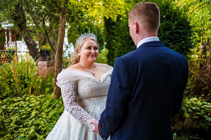 A bride in a white lace wedding gown and tiara holds hands with a groom in a navy suit. They are standing outdoors in a lush garden with green foliage and trees in the background. The bride is smiling at the groom. Image by Documentary Wedding Photographer Lewis Bishop Photography.