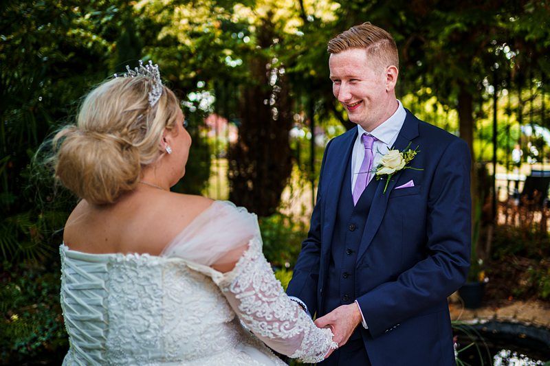 A bride and groom stand outdoors holding hands. The bride, in a white lace dress and tiara, faces the groom, who is in a navy blue suit with a lavender tie and boutonnière. They are smiling at each other. Trees and greenery surround them in the background. Image by Documentary Wedding Photographer Lewis Bishop Photography.