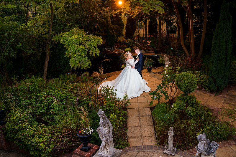 A bride and groom share an intimate dance in an elegantly landscaped garden at night. The bride wears a flowing white gown, and the groom is in a dark suit. They are surrounded by greenery, statues, and a softly illuminated pond in the background. Image by Documentary Wedding Photographer Lewis Bishop Photography.
