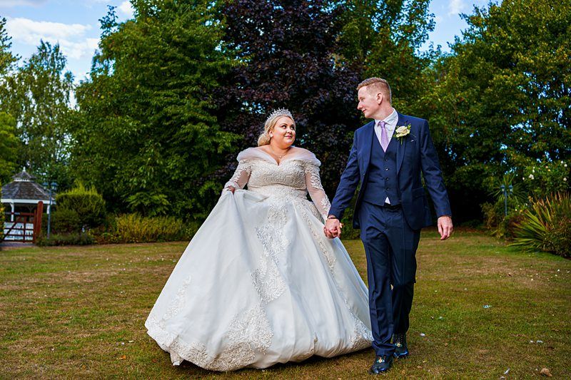 A couple walks hand in hand outdoors on their wedding day. The bride wears a white lace gown and the groom is in a dark suit with a vest and tie. They are smiling at each other with trees and a garden in the background. Image by Documentary Wedding Photographer Lewis Bishop Photography.