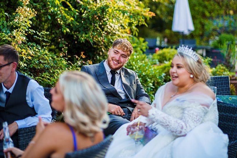 A bride in a white dress and tiara, and a groom in a grey suit sit outdoors with friends. They are smiling and having a conversation. The background is filled with lush greenery and patio furniture. Image by Documentary Wedding Photographer Lewis Bishop Photography.