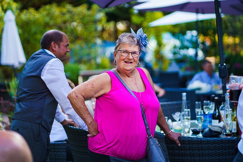 An older woman wearing a bright pink top and a blue feathered headpiece is smiling as she poses outdoors. A man in a white shirt and grey vest stands nearby. The setting appears to be a social gathering with tables, drinks, and people in the background. Image by Documentary Wedding Photographer Lewis Bishop Photography.