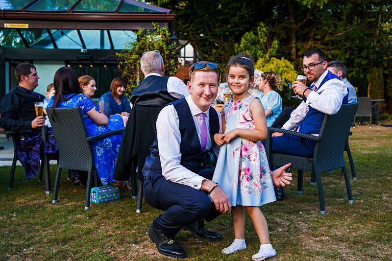 A man squats down and smiles for a photo with a young girl in a light blue dress. They are outdoors at an event with other people sitting and chatting in the background. Some attendees hold drinks, and greenery surrounds the area. Image by Documentary Wedding Photographer Lewis Bishop Photography.