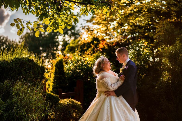 A bride and groom stand in a lush garden, illuminated by warm sunlight. The bride wears a white lace gown and the groom is in a dark suit. They look lovingly into each other's eyes, surrounded by green foliage and trees under a clear sky. Image by Documentary Wedding Photographer Lewis Bishop Photography.
