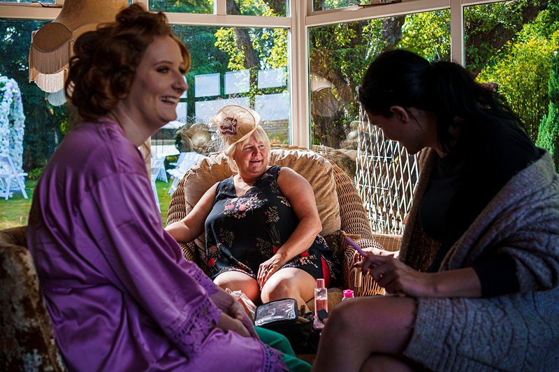 Three women are sitting in a sunlit room, enjoying a conversation. One woman, wearing a purple robe, is smiling, another, seated in a wicker chair, is laughing, and the third, dressed in dark clothes, is speaking while holding a makeup product. The outside garden is visible. Image by Documentary Wedding Photographer Lewis Bishop Photography.
