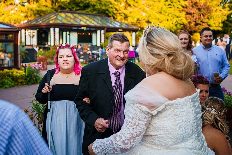 A man in a dark suit and pink tie smiles warmly at a woman in a white wedding dress and crown. A person with pink hair and a black and gray off-the-shoulder dress stands nearby, holding a microphone. Several onlookers stand in the background in an outdoor setting. Image by Documentary Wedding Photographer Lewis Bishop Photography.