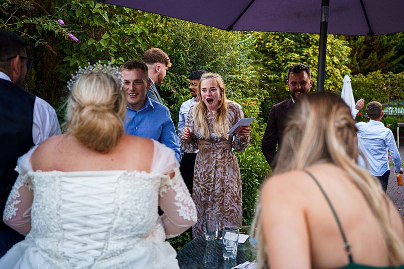 A bride in a white wedding dress and guests in various attire are gathered outdoors under an umbrella. One woman appears excited and is holding a piece of paper. There are trees and greenery in the background. Image by Documentary Wedding Photographer Lewis Bishop Photography.