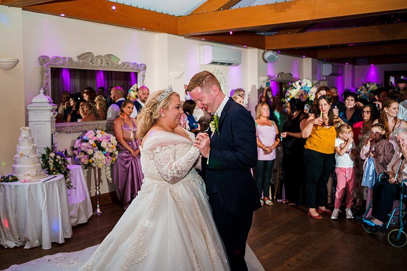 A bride and groom share their first dance at their wedding reception. The bride wears a white gown and a tiara, while the groom is in a dark suit. Guests surround them, watching and smiling. A decorated wedding cake and floral arrangements are visible in the background. Image by Documentary Wedding Photographer Lewis Bishop Photography.