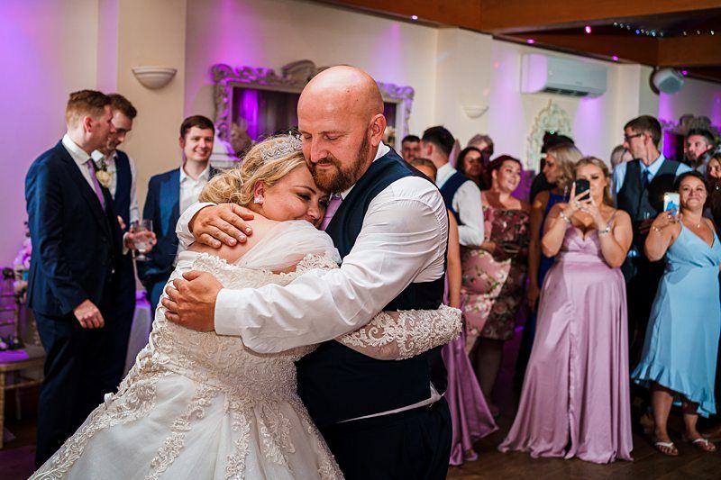 A bride and groom embrace during their wedding reception, surrounded by guests. The bride is in a white lace gown and the groom wears a vest and tie. Guests in formal attire look on, some taking photos with their phones. The room is lit with purple lights. Image by Documentary Wedding Photographer Lewis Bishop Photography.