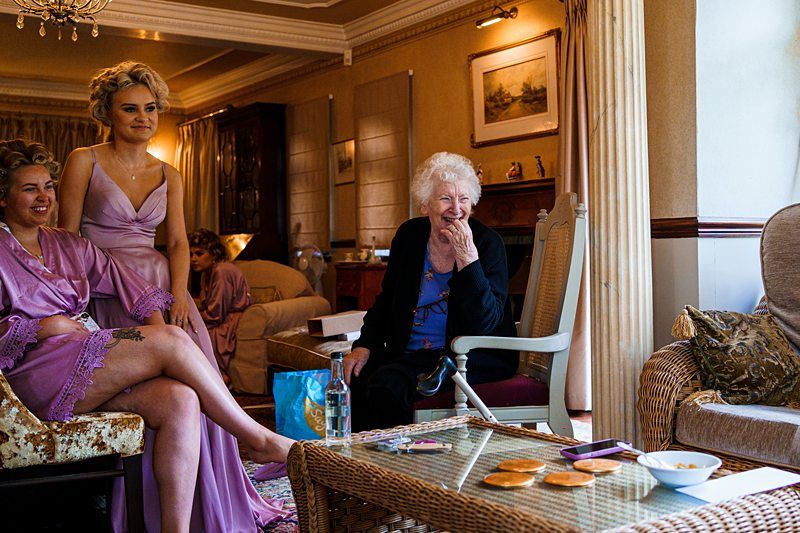 A joyful scene in a living room with a smiling elderly woman sitting on a chair and two women in matching pink robes, one of whom has a tattoo on her thigh. Snacks and drinks rest on a wicker table, suggesting a relaxed gathering or celebration. Image by Documentary Wedding Photographer Lewis Bishop Photography.