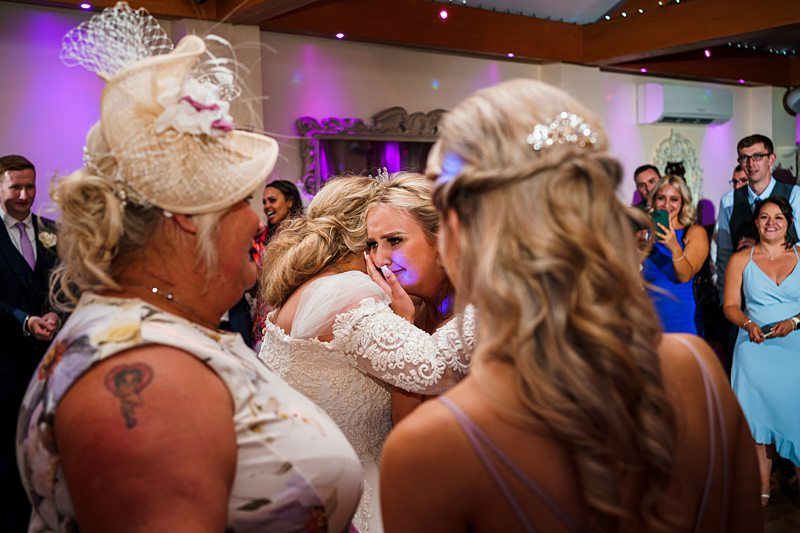 A bride hugs a woman while shedding tears at a wedding reception. The bride is in a white lace dress, and the woman she's hugging has blonde hair. Surrounding them are guests watching, with one prominently in a floral dress and a beige fascinator. Image by Documentary Wedding Photographer Lewis Bishop Photography.