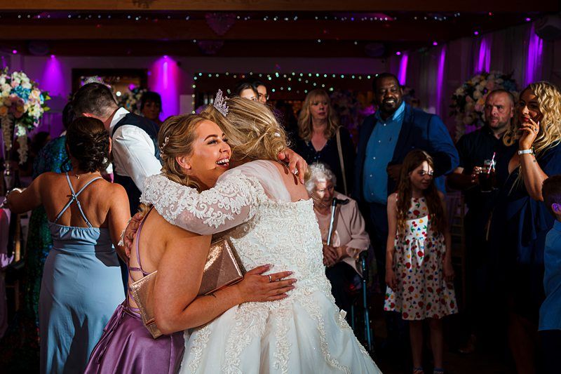 A bride in a white gown and veil embraces a bridesmaid wearing a lilac dress at a wedding reception. Guests in colorful outfits stand around them, some smiling and others talking, with a backdrop of purple lighting and floral decorations. Image by Documentary Wedding Photographer Lewis Bishop Photography.