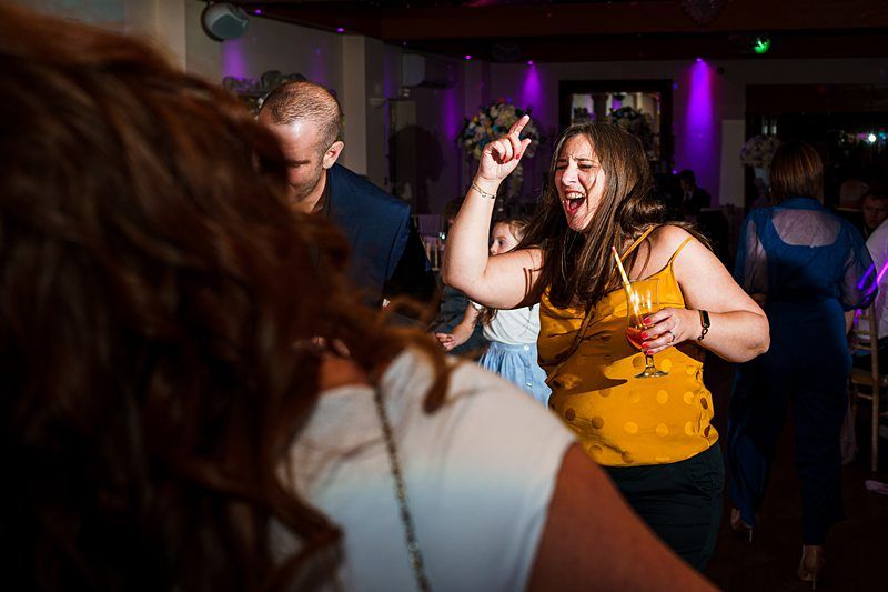 A woman in a yellow polka dot sleeveless top holds a drink in one hand and points upward with the other while dancing at a party. She appears to be singing or shouting joyfully. Other guests are around her, and the room is illuminated with colorful lighting. Image by Documentary Wedding Photographer Lewis Bishop Photography.
