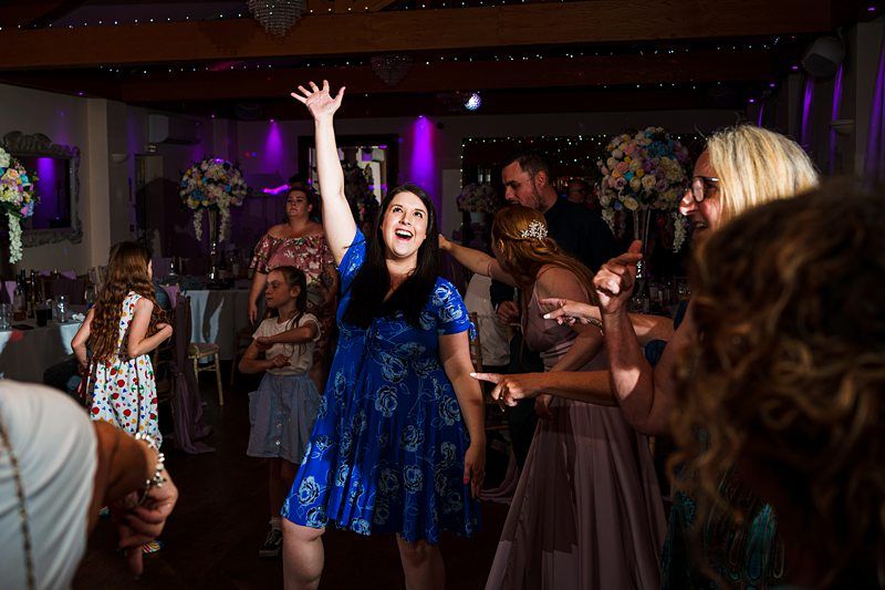 A woman in a blue dress joyfully raises her hand while dancing at a lively event. Other attendees, including children and adults, also dance and socialize in the background. The room is decorated with flowers and lit with purple and white lights. Image by Documentary Wedding Photographer Lewis Bishop Photography.