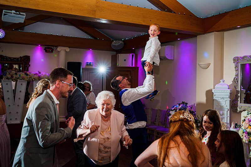 A group of people are dancing and having fun at a party. A man lifts a smiling child on his shoulders, while others, including an elderly woman and a man in a suit, join in the festivities. The room is illuminated with colorful lights and decorated with flowers. Image by Documentary Wedding Photographer Lewis Bishop Photography.